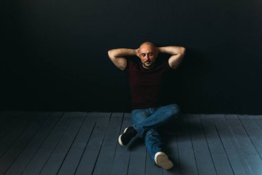 Gözlüklü, kot pantolonlu, kel, yerde, siyah bir duvara yaslanmış genç bir adam.