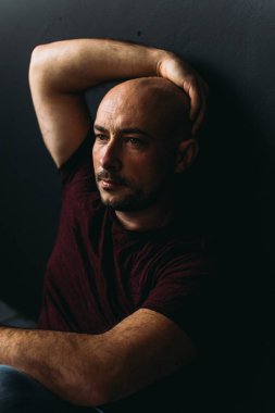 Gözlüklü, kot pantolonlu, kel, yerde, siyah bir duvara yaslanmış genç bir adam.