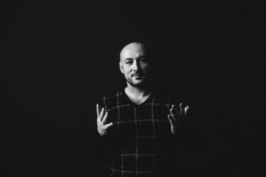 Gözlüklü, koyu renk elbiseli, kel, siyah arka planda genç bir adam elleri ve yüzünde duygular olan bir şey gösterir.