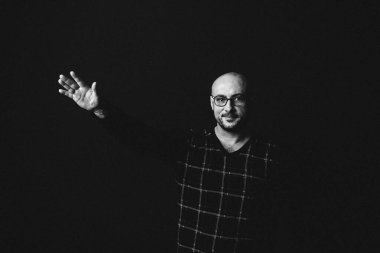 Gözlüklü, koyu renk elbiseli, kel, siyah arka planda genç bir adam elleri ve yüzünde duygular olan bir şey gösterir.