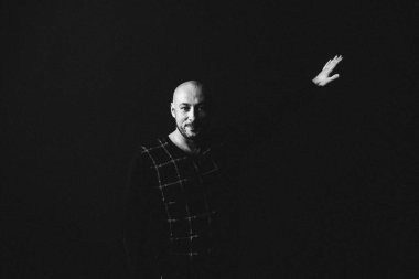 Gözlüklü, koyu renk elbiseli, kel, siyah arka planda genç bir adam elleri ve yüzünde duygular olan bir şey gösterir.