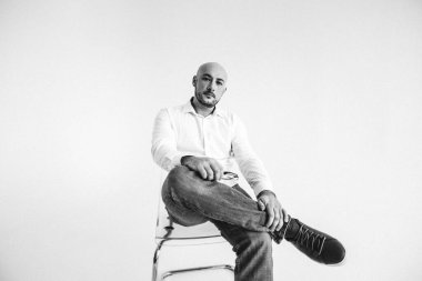 Gözlüklü genç bir adam, beyaz gömlekli, züppe, kel, beyaz arka planda bir sandalyeye oturmuş, elleri yüzünde duygularla bir şeyler gösteriyor.