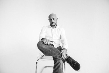 Gözlüklü genç bir adam, beyaz gömlekli, züppe, kel, beyaz arka planda bir sandalyeye oturmuş, elleri yüzünde duygularla bir şeyler gösteriyor.