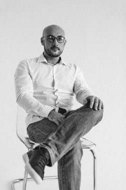 Gözlüklü genç bir adam, beyaz gömlekli, züppe, kel, beyaz arka planda bir sandalyeye oturmuş, elleri yüzünde duygularla bir şeyler gösteriyor.