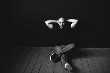 Gözlüklü, kot pantolonlu, kel, yerde, siyah bir duvara yaslanmış genç bir adam.