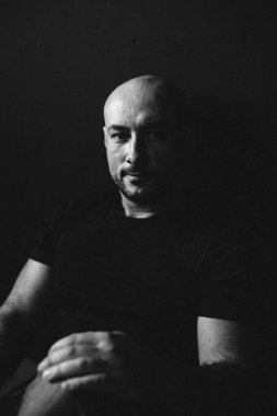 Gözlüklü, kot pantolonlu, kel, yerde, siyah bir duvara yaslanmış genç bir adam.