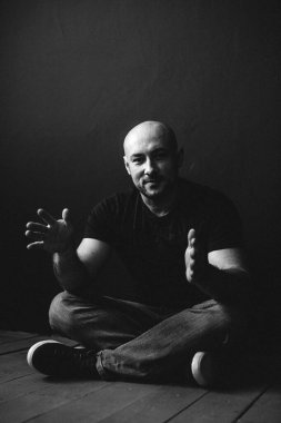 Gözlüklü, kot pantolonlu, kel, yerde, siyah bir duvara yaslanmış genç bir adam.