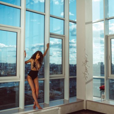 Güzel vücut şekline sahip genç bir kız, güzel iç çamaşırları ve çıplaklığıyla, akşam vakti panoramik pencereli bir odada, akşam güneşi üzerine düşüyor.