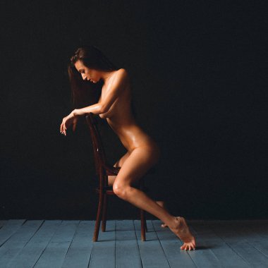 Güzel vücutlu genç bir kız, çıplak, siyah bir duvara yaslanmış, eski bir sandalyeye oturuyor, vücudu yağla ovulmuş.