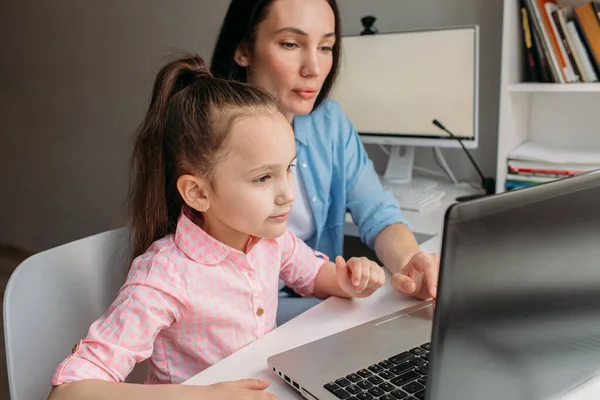 Mom and daughter are at home today. They work remotely on a laptop. Mom helps her daughter complete tasks on a computer.