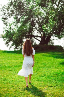 Beyaz yaz elbisesi içinde genç güzel bir kız gölde yeşil bir çayırda yürüyor, yaz güneşi yeşillikleri içinde.