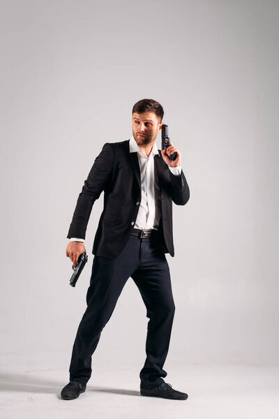 A young guy in a black suit with a gun in his hands on a white background in the studio, he depicts a security guard, bodyguard or Agent