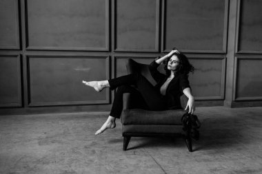 Siyah takım elbiseli güzel kıvırcık kız çıplak bir vücutta, stüdyoda bir koltuk, bir tezgah ve gri bir duvar, siyah beyaz bir fotoğraf