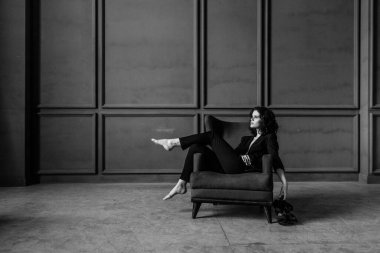 Siyah takım elbiseli güzel kıvırcık kız çıplak bir vücutta, stüdyoda bir koltuk, bir tezgah ve gri bir duvar, siyah beyaz bir fotoğraf