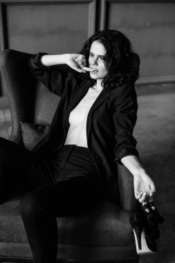 Siyah takım elbiseli güzel kıvırcık kız çıplak bir vücutta, stüdyoda bir koltuk, bir tezgah ve gri bir duvar, siyah beyaz bir fotoğraf