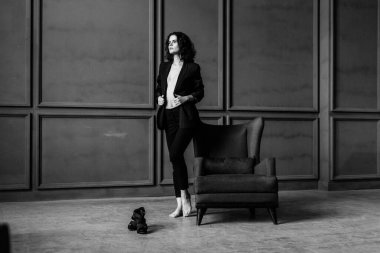 Siyah takım elbiseli güzel kıvırcık kız çıplak bir vücutta, stüdyoda bir koltuk, bir tezgah ve gri bir duvar, siyah beyaz bir fotoğraf