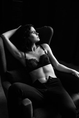 Siyah takım elbiseli güzel kıvırcık kız çıplak bir vücutta, stüdyoda bir koltuk, bir tezgah ve gri bir duvar, siyah beyaz bir fotoğraf