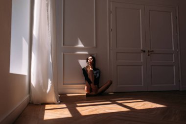 Genç, güzel kız model, uzun boylu, siyah vücutlu, büyük, parlak bir odada, pencereden gelen parlak güneş ışığı üzerine düşüyor.