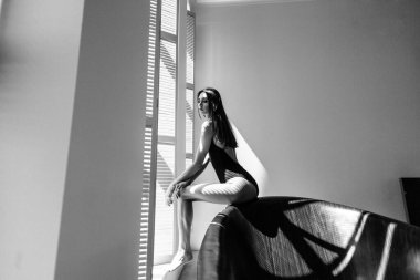 Genç, güzel kız model, uzun boylu, siyah vücutlu, büyük, parlak bir odada, pencereden gelen parlak güneş ışığı üzerine düşüyor.