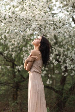 Kıvırcık saçlı genç bir kız, gün batımında, çiçek açan ağaçların arasında, bir elma bahçesinde, gün ışığında duruyor.