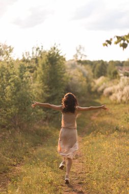 Kıvırcık saçlı genç bir kız, gün batımında, çiçek açan ağaçların arasında, gün ışığında duruyor.