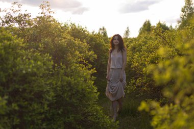 Akşamları kıvırcık saçlı genç bir kız. Gün batımında yeşil ağaçların arasında, basit bir elbiseyle gün ışığında duruyor.