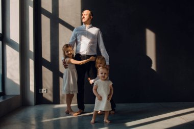 Baba ve çocukları karanlık duvarları olan güneşli bir odada babalarını seviyorlar, onunla oynuyorlar ve sarılıyorlar.