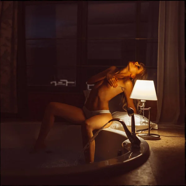 young beautiful blonde girl, evening in the hotel room alone in white lingerie and half-naked under the light from the lamp