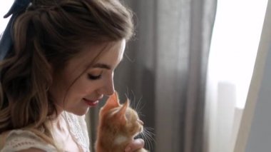 Tarafsız elbiseli güzel bir kız pencerenin yanındaki evde bir resim çizer ve küçük kızıl saçlı bir kedi yavrusuyla oynar. Kedi yavrusu fırçayla oynar.