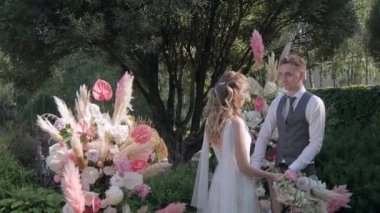 Gelin ve damat, taze çiçeklerin düğün kemerinin arka planında durup birbirlerine aşkla bakıyorlar.