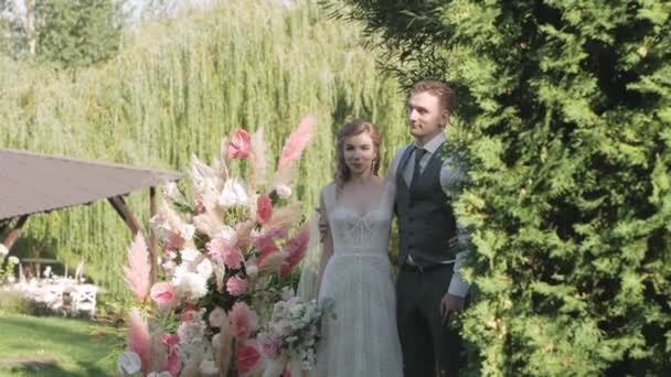 夏天的一天 新郎和新娘站在婚礼拱门的后面 那拱门由鲜花和蓬松的青草做成 中等大小 面带微笑 — 图库视频影像