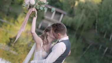 Ahşap bir köprüde damat, yeşil bir gölün ve ağaçların arka planında gelini gözleri kapalı öper, kız bir buket taze çiçeği kaldırır.