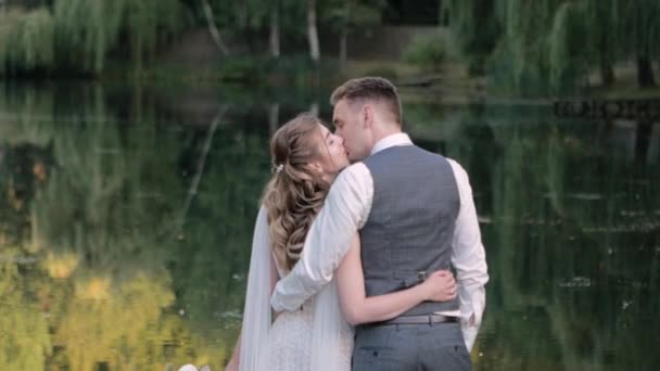 白い花嫁と新郎が木製の橋の上に立って 夏の日に 彼らは緑の湖の背景に抱擁し 抱擁は彼らがカメラに背を向けて立つ彼女にキスをする — ストック動画