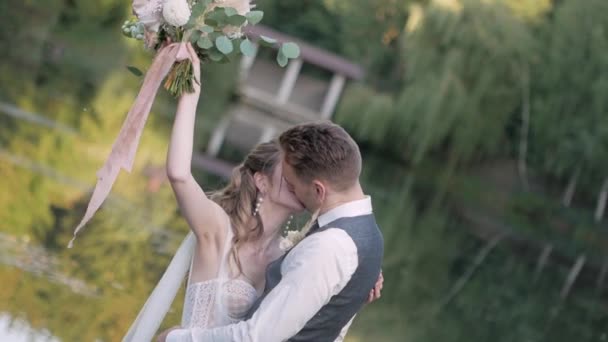 新郎在一座木桥上 靠着绿湖和树木 闭上眼睛吻着新娘 姑娘举起一束鲜花 — 图库视频影像