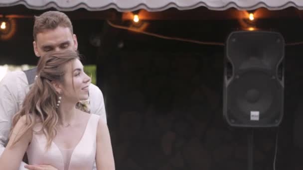 Un jeune couple danse joyeusement et tendrement sur le fond d'un gazebo en bois et des ampoules, le marié embrasse doucement la mariée par derrière, ils sourient et sont heureux le ciel ouvert 