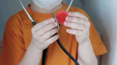 Maskeli, kahverengi saçlı, turuncu tişörtlü merdivenlerdeki evde oturan ve virüse benzeyen bir stetoskobu olan bir oyuncağı dinleyen bir çocuk.