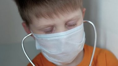 Beyaz arka planda, turuncu tişörtlü, tıbbi maskeli bir çocuk steteskopla bir şey dinliyor ve dikkatlice altına bakıyor.