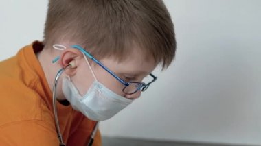 Dairesinde, odasında, turuncu tişörtlü, gözlüklü, yerde güzel bir battaniyeyle oturan beyaz arka planda steteskoplu oyuncakları dinleyen ve ciddi ciddi düşünen bir çocuk..