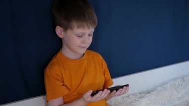 Sarı tişörtlü bir çocuk, kahverengi saçlı, battaniyeyle yapılmış bir yatakta oturuyor, yeşil arka planda, elinde bir telefonla, çizgi film izliyor, ve odada, kamerada komik suratlar yapıyor..