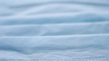 Mavi renkli mikroplardan korunmak için tıbbi maske beyaz bir yüzeyde gündüz güneşinin yakın çekiminde bulunur. Kumaşın dokusu görünür. Hareketsiz durağan bir çerçeve.