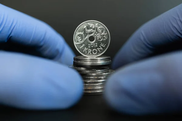 Norwegian financial crisis, health system and economy during coronavirus or covid-19 pandemic. One Norwegian krone coin standing on stack of coins surronded by hands with protective gloves.