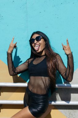 Uzun saçlı ve güneş gözlüklü esmer bir kızın dikey fotoğrafı. Manken kılığına girmiş ve ellerini kaldırarak rock 'n roll' a atfedilen hareketi yapıyor.