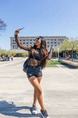 Uzun saçlı, güneş gözlüklü ve yaz örgüsü elbiseli esmer bir kızın dikey fotoğrafı. Dilini sokağın ortasında dışarı çıkararak selfie çekiyor.