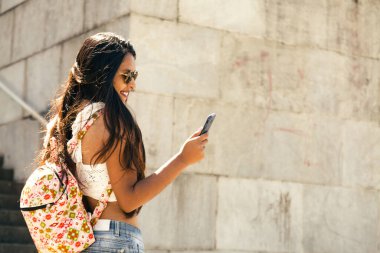 Güneş gözlüklü ve yazlık elbiseli esmer bir kızın profili. Bir elinde cep telefonu, diğer elinde kitap var. Bir merdivenin ortasında antik bir binanın önünde duruyor.