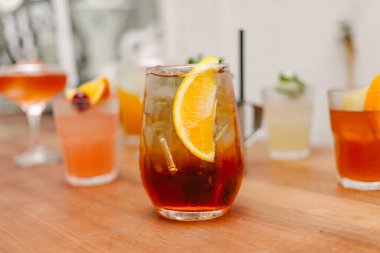 Yanında diğer kokteyllerle birlikte ahşap bir masada süslemek için bir parça portakal içeren buzlu bir kristal bardak.