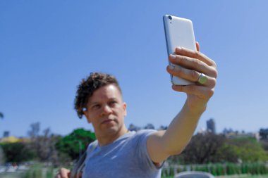Kıvırcık saçlı, modern saçlı, yüzük takan ve cep telefonuyla parkın ortasında selfie çeken bir adam.