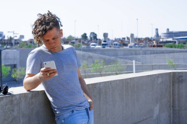 Kıvırcık saçlı bir adam cep telefonunu kullanarak arka planda şehir olan bir duvara yaslanıyor.