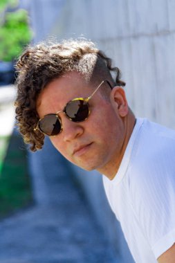 Kıvırcık saçlı, güneş gözlüklü, beton duvara yaslanmış ve yüzünü yana çevirmiş şık bir adamın portresi.