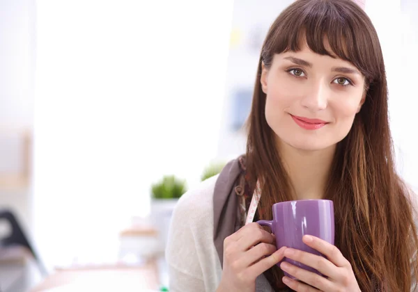 Young attractive fashion designer standing by desk in office, holding cup — Stock Photo, Image