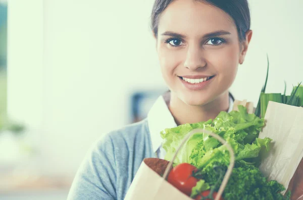 Mujer joven sosteniendo bolsa de la compra de comestibles con verduras. De pie en la cocina —  Fotos de Stock